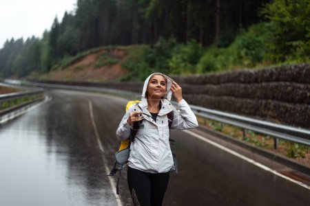 Téléchargez les photos : Portrait de jeune femme en imperméable debout sur route asphaltée humide et souriante. Charme voyageur randonnée à la montagne pendant les loisirs. - en image libre de droit
