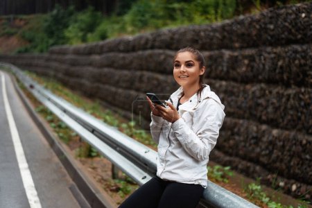 Téléchargez les photos : Touriste souriante utilisant un téléphone portable moderne à l'extérieur. Heureuse femme caucasienne randonnée avec sac à dos parmi les montagnes. Concept de personnes et de technologie. - en image libre de droit