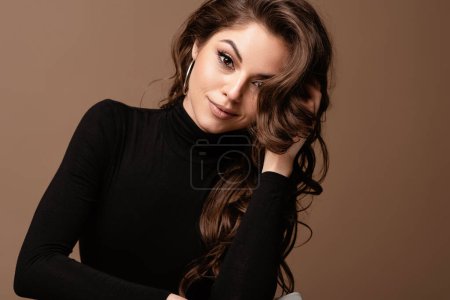 Photo for Beautiful smiling young european woman with seductive look portrait. Girl wear black sweater with beautiful face and hair sit on chair and looking at camera. Isolated on brown background. Copy space - Royalty Free Image