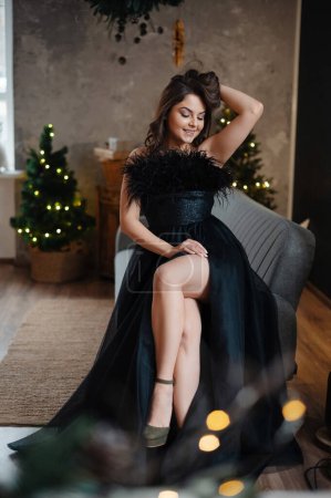 Foto de Hermosa chica en vestido de noche sentada en el sillón cerca del árbol de Navidad. Fondo interior de lujo. Vacaciones de Navidad, celebración, concepto de regalos - Imagen libre de derechos