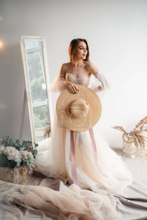 Téléchargez les photos : Vertical view of the bride in wedding dress standing in front of the window with hat at the hand. Romantic atmosphere of the bride's morning concept - en image libre de droit