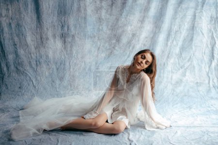 Foto de Full length view of the young brunette bride in white luxury wedding dress. Loft studio interior with blue fabric background. Looking down - Imagen libre de derechos