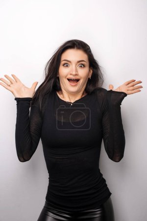 Foto de Portrait of shocked young brunette woman in dark clothes standing and screaming at camera with surprised face. Indoor studio shot isolated on light white background - Imagen libre de derechos