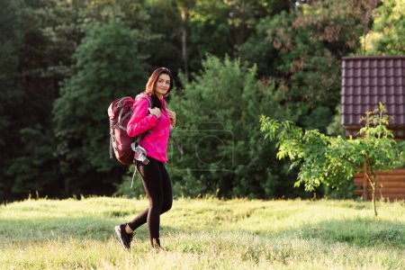 Photo for Young european woman walking with backpack on green meadow near forest. Beautiful smiling girl wear sportswear and pink jacket. Concept of resting and tourism on nature. Sunny daytime - Royalty Free Image