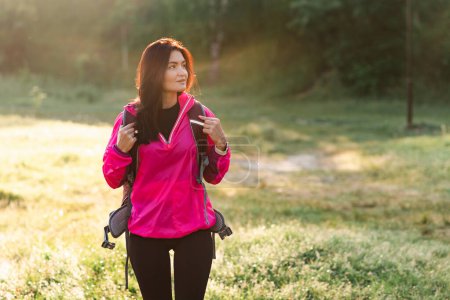 Foto de Joven mujer europea caminando con mochila en prado verde cerca del bosque. Atractiva chica enfocada a usar ropa deportiva y chaqueta rosa. Concepto de descanso y turismo en la naturaleza. Día soleado - Imagen libre de derechos