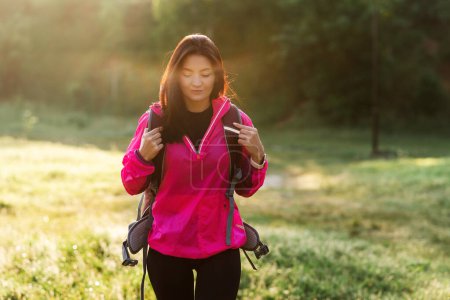 Photo for Young european woman walking with backpack on green meadow near forest. Attractive focused girl wear sportswear and pink jacket. Concept of resting and tourism on nature. Sunny daytime - Royalty Free Image
