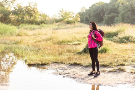 Photo for Young european woman standing with backpack on lake coast and enjoying view. Beautiful smiling girl wear sportswear and pink jacket. Concept of resting and tourism on nature. Sunny daytime - Royalty Free Image