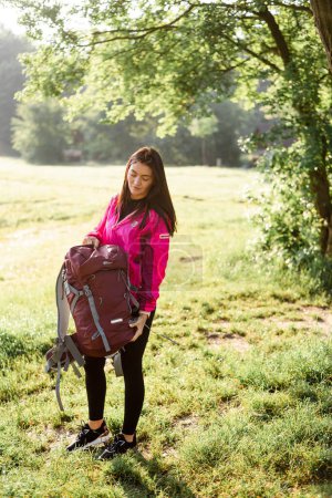 Téléchargez les photos : Jeune femme européenne ouvrant son sac à dos sur prairie verte près de la forêt. Belle fille souriante porter des vêtements de sport et veste rose. Concept de repos et de tourisme sur la nature. Journée ensoleillée - en image libre de droit
