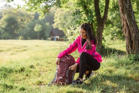 Photo for Young european woman opening her backpack on green meadow near forest. Beautiful smiling girl wear sportswear and pink jacket. Concept of resting and tourism on nature. Sunny daytime - Royalty Free Image