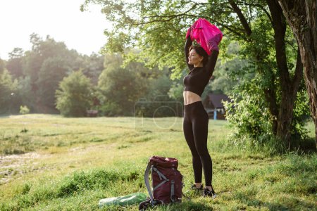Photo for European athlete girl take off pink jacket on green meadow near forest. Young beautiful smiling woman with closed eyes wear sportswear. Concept of resting and tourism on nature. Sunny daytime - Royalty Free Image