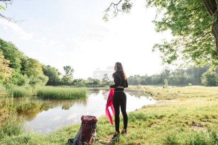 Téléchargez les photos : Une athlète européenne enlève sa veste rose sur un pré vert près de la forêt. Jeune belle femme souriante aux yeux fermés porter des vêtements de sport. Concept de repos et de tourisme sur la nature. Journée ensoleillée - en image libre de droit
