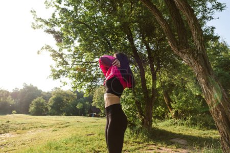 Photo for European athlete girl take off pink jacket on green meadow near forest. Young beautiful smiling woman with closed eyes wear sportswear. Concept of resting and tourism on nature. Sunny daytime - Royalty Free Image