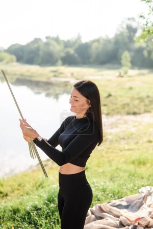 Foto de European athlete girl holding aluminum flexible arcs-sticks for tent. Young pretty smiling woman standing on green meadow and wear sportswear. Concept of resting and tourism on nature. Sunny daytime - Imagen libre de derechos