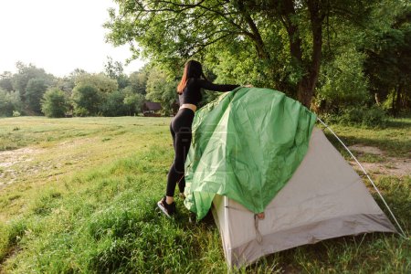 Photo for Athlete girl covering outer tent canvas on tent. Green meadow near forest. Back view of sexy woman wear sportswear. Concept of resting and tourism on nature. Camping vacation. Sunny summer daytime - Royalty Free Image