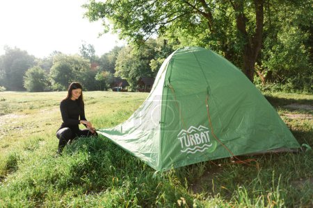 Foto de Athlete girl covering outer tent canvas on tent. Green meadow near forest. Back view of sexy woman wear sportswear. Concept of resting and tourism on nature. Camping vacation. Sunny summer daytime - Imagen libre de derechos