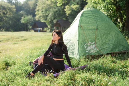 Photo for European sport girl sitting on fitness mat near tent and enjoying sunlight. Green meadow. Young beautiful woman wear sportswear. Concept of resting and tourism on nature. Camping vacation. Sunny day - Royalty Free Image
