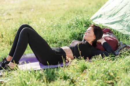 Photo for European sport girl lying on fitness mat near tent and enjoying sunlight. Green meadow. Young beautiful woman wear sportswear. Concept of resting and tourism on nature. Camping vacation. Sunny day - Royalty Free Image