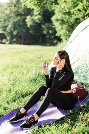 Téléchargez les photos : Européen fille sportive assis sur tapis de fitness près de tente et boire du thé. Prairie verte près du lac. Jeune jolie femme porter des vêtements de sport et profiter de la vue. Repos et tourisme sur la nature. Vacances en camping. Journée ensoleillée - en image libre de droit