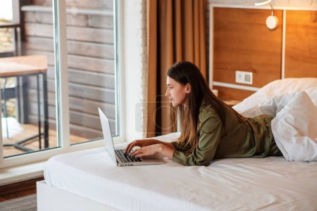 Photo for Beautiful european girl lying on bed and using laptop computer. Side view of young smiling woman with brown hair wear pajamas. Concept of rest. Interior of bedroom in modern apartment. Sunny daytime - Royalty Free Image