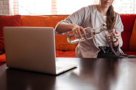 Foto de Girl sitting on sofa at table with laptop and pouring water to glass at home. Obscure face of young woman with brown hair wear casual clothes. Interior of living room in modern apartment - Imagen libre de derechos