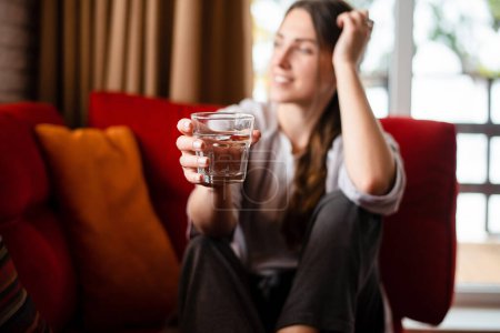 Photo for Focus on foreground of glass with water in hand of girl. Blurred view of young european smiling woman wear casual clothes. Interior of living room in modern apartment. Daytime - Royalty Free Image