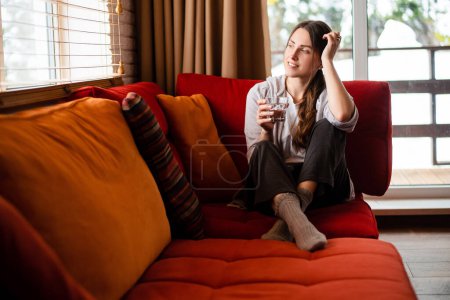 Photo for European smiling girl sit on sofa and drinking water from glass at home. Young thoughtful woman wear casual clothes. Interior of living room in modern apartment. Daytime - Royalty Free Image