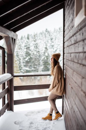 Photo for Charming young woman standing on balcony of modern wooden cottage and enjoying view on snowy forest. Beautiful winter weather. Travelling concept. - Royalty Free Image