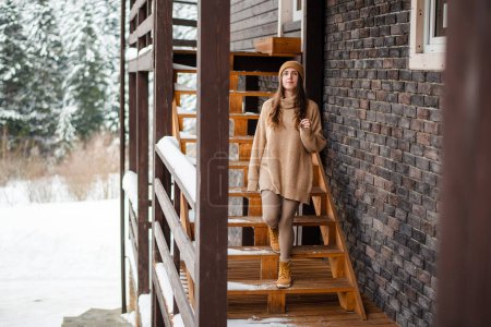 Téléchargez les photos : Belle femme caucasienne marchant sur les escaliers en bois du chalet moderne et regardant la nature enneigée autour. Vacances d'hiver dans les montagnes. Concept de modes de vie. - en image libre de droit