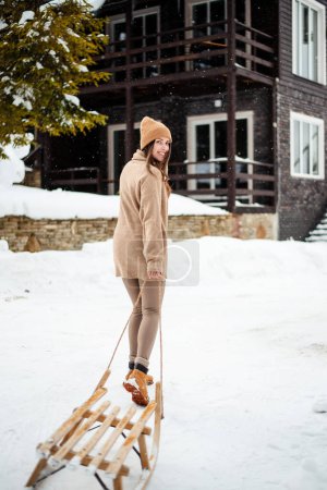 Photo for Cheerful caucasian woman smiling on camera while walking with empty wooden sledge outdoors. Winter season for active time spending. Beautiful snowy nature around. - Royalty Free Image