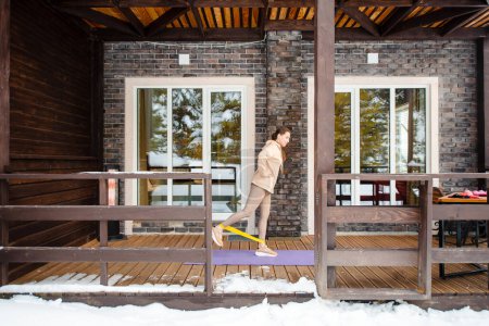 Photo for Active caucasian woman using rubber band for training legs on terrace of modern wooden cottage. Snow lying around. Sport activity during winter holidays. - Royalty Free Image