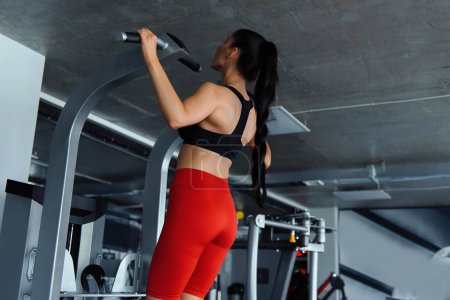 Photo for Back view of young woman with muscular body in black bra and red shorts training arms on gym machine. Strong brunette exercising regularly for staying fit and healthy. - Royalty Free Image