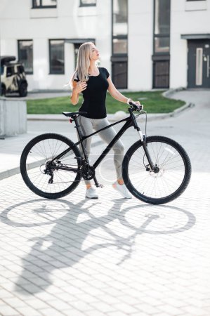 Photo for Beautiful caucasian woman standing outdoors with black bicycle and keeping eyes closed. Young female with blond hair in sport clothes spending free time for training on fresh air. - Royalty Free Image