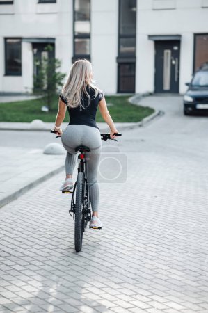 Foto de Vista posterior de la mujer joven vestida con ropa deportiva montar en bicicleta en la calle de la ciudad. Rubia caucásica activa pasando la mañana para el ciclismo. Concepto de estilos de vida saludables. - Imagen libre de derechos