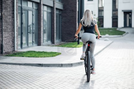 Photo for Back view of young woman dressed in sport clothes riding bike on city street. Active caucasian blonde spending morning time for cycling. Healthy lifestyles concept. - Royalty Free Image