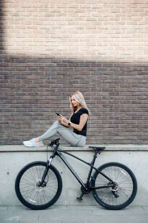 Foto de Mujer atractiva con forma de cuerpo atlético sentado al aire libre cerca de la bicicleta negra y el uso de un teléfono inteligente moderno. Mujer caucásica en ropa deportiva tomando un descanso después del entrenamiento al aire libre. - Imagen libre de derechos