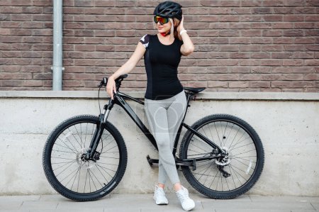 Téléchargez les photos : Portrait complet de femme sportive en vêtements de sport, casque et lunettes debout avec vélo noir dans la rue de la ville. Concept de personnes, de cyclisme et de loisirs. - en image libre de droit