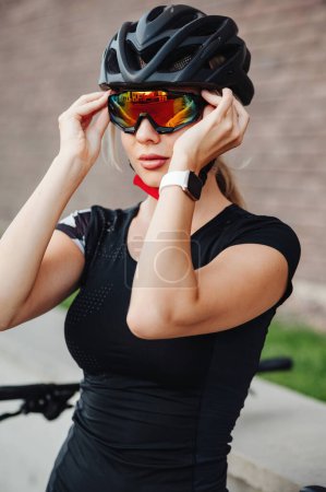 Téléchargez les photos : Fitness jeune femme en casque de protection et lunettes de sport posant sur la rue de la ville. Dame caucasienne avec montre intelligente au poignet se préparant pour le cyclisme matinal sur l'air frais. - en image libre de droit