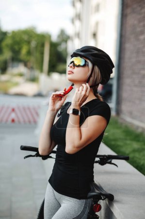 Photo for Side view of pretty young woman wearing safety helmet and mirrored glasses while standing on street with black bike. Female cyclist preparing for outdoors workout. - Royalty Free Image