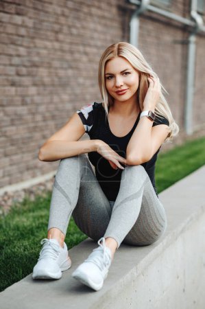 Photo for Active young woman wearing sport clothes and smart watch on wrist resting after cycling on fresh air. Healthy and fit blonde sitting on city street and looking down. - Royalty Free Image