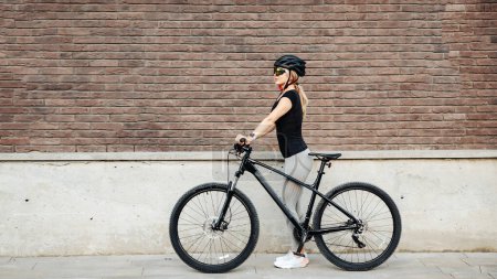 Téléchargez les photos : Vue latérale de cycliste caucasienne en vêtements de sport, casque et lunettes debout avec vélo noir sur la rue de la ville. Détente après l'entraînement. - en image libre de droit