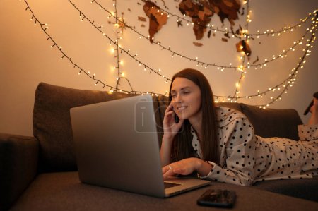 Photo for Woman laying at the sofa chatting with friends on modern laptop. Positive lady resting at modern apartment, having fun and relax at evening - Royalty Free Image