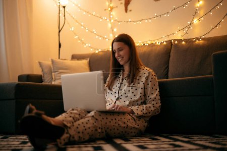 Photo for Happy young woman sitting near the coach browsing internet chatting with friends on modern laptop. Positive lady resting at modern apartment, having fun and relax at evening - Royalty Free Image