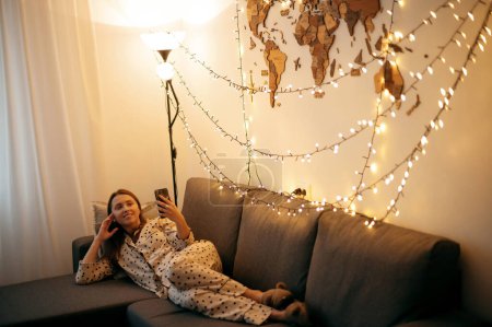 Foto de Hermosa chica usando un teléfono inteligente mientras está acostada en el sofá en casa, su piso iluminado con luz de guirnalda. Mujer linda sonríe y navegar a través de Internet - Imagen libre de derechos