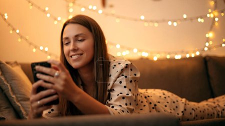 Téléchargez les photos : Femme heureuse utilisant smartphone le soir à la maison tout en étant allongé sur le canapé. Fille souriante en pyjama relaxant à la chambre décorée de Noël - en image libre de droit