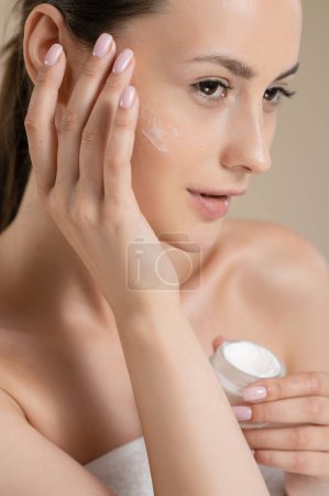 Photo for Sensual young woman with bare shoulders applying cream on face in studio. Caucasian female model enjoying beauty rituals over beige background. Fresh natural skin. - Royalty Free Image