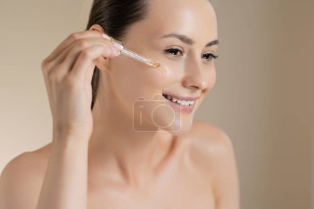 Photo for Charming dark haired woman applying serum on face from pipette. Young lady with naked shoulders using cosmetic for beauty procedures. isolated over beige studio background. - Royalty Free Image