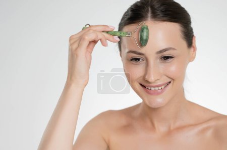 Photo for Happy young brunette with beauty roller in hands lifting skin on fresh face. Caucasian female model enjoying daily rituals. Isolated over white studio background. - Royalty Free Image