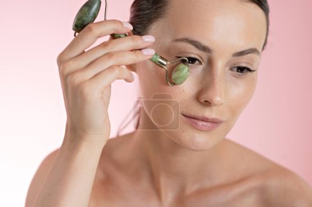 Photo for Pretty caucasian woman massaging skin under eyes with beauty roller over pink background. Female model doing face procedures for lifting effect. Anti-aging rituals. - Royalty Free Image