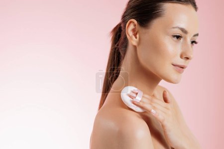 Photo for Stunning young woman with bare shoulder using soft sponge for applying powder on cheeks. Caucasian female model doing light makeup. Isolated over pink studio background. - Royalty Free Image