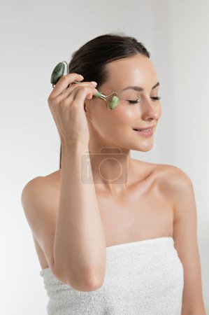 Photo for Pleasant caucasian model with natural beauty doing skin procedures with jade roller. Young woman with brown hair wrapped in towel. Isolated over white studio background. - Royalty Free Image